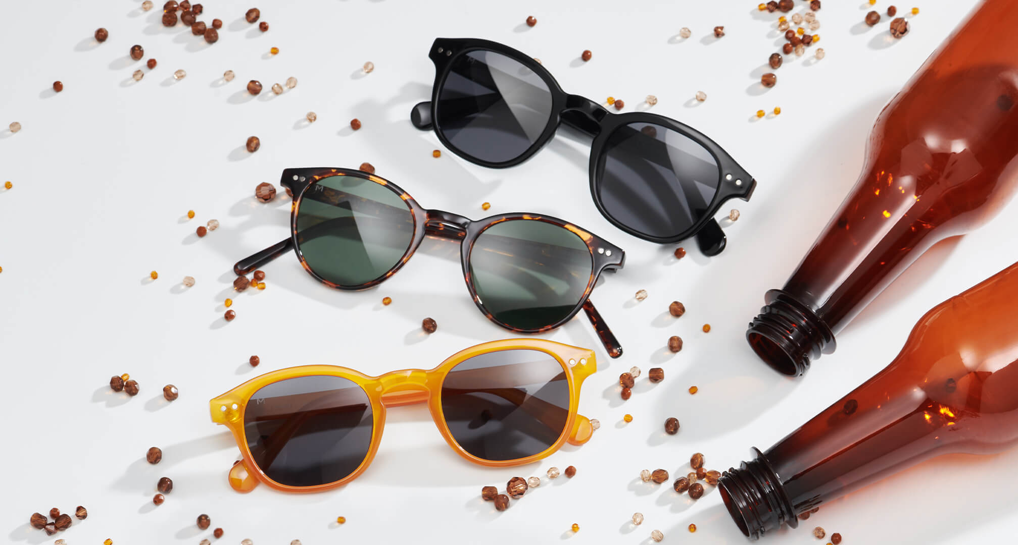 trzy pary okularów przeciwsłonecznych wykonanych z trwałych materiałów na białym stole, z koralikami i plastikowymi butelkami wokół nich
